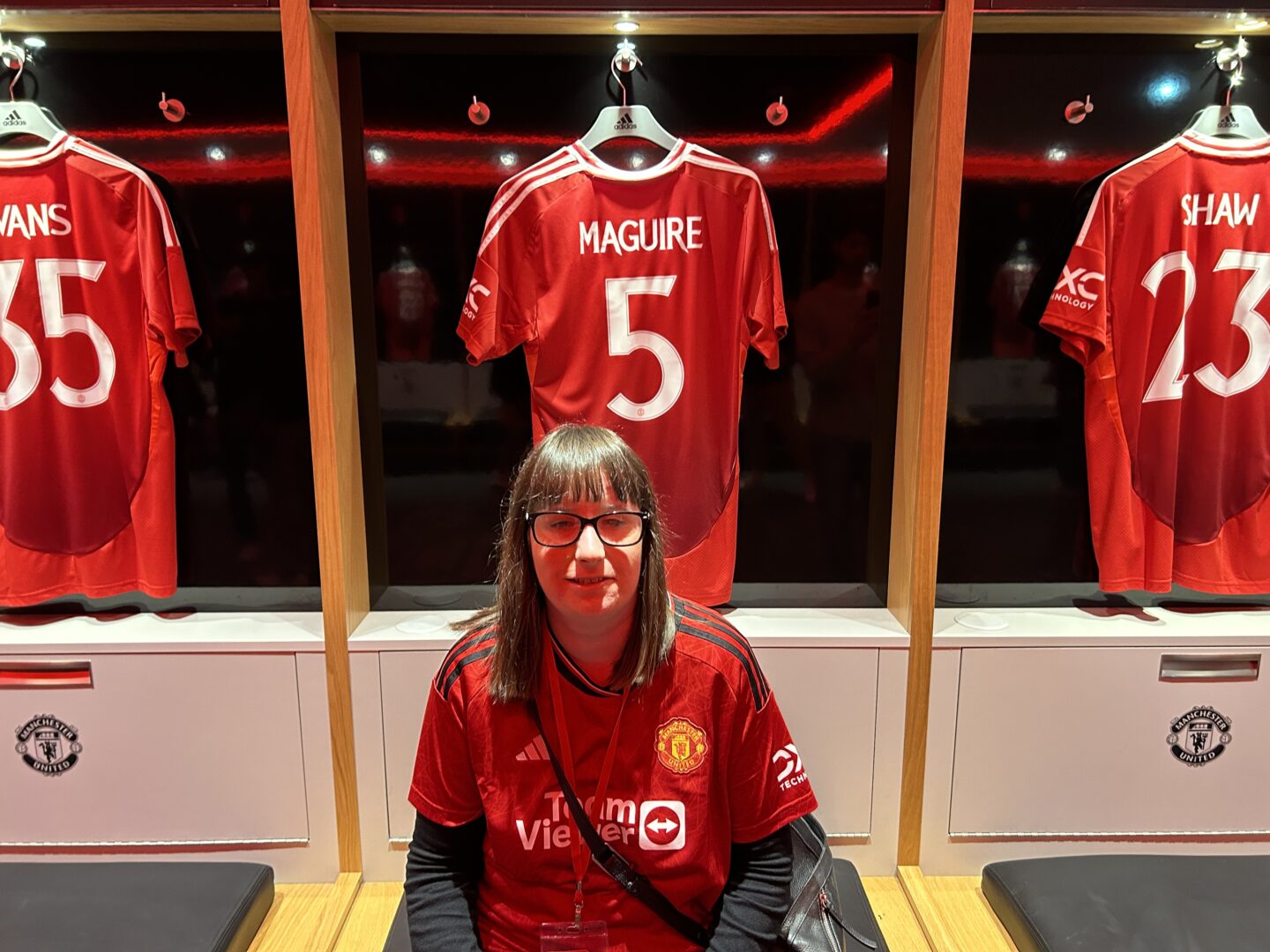 Holly sat in the players changing room at Old Trafford. She has shoulder-length brown hair and is wearing a Manchester United shirt and glasses. There are three Manchester United shirts hanging in lockers behind her: Evans - 35, Maguire – 5, and Shaw - 23.