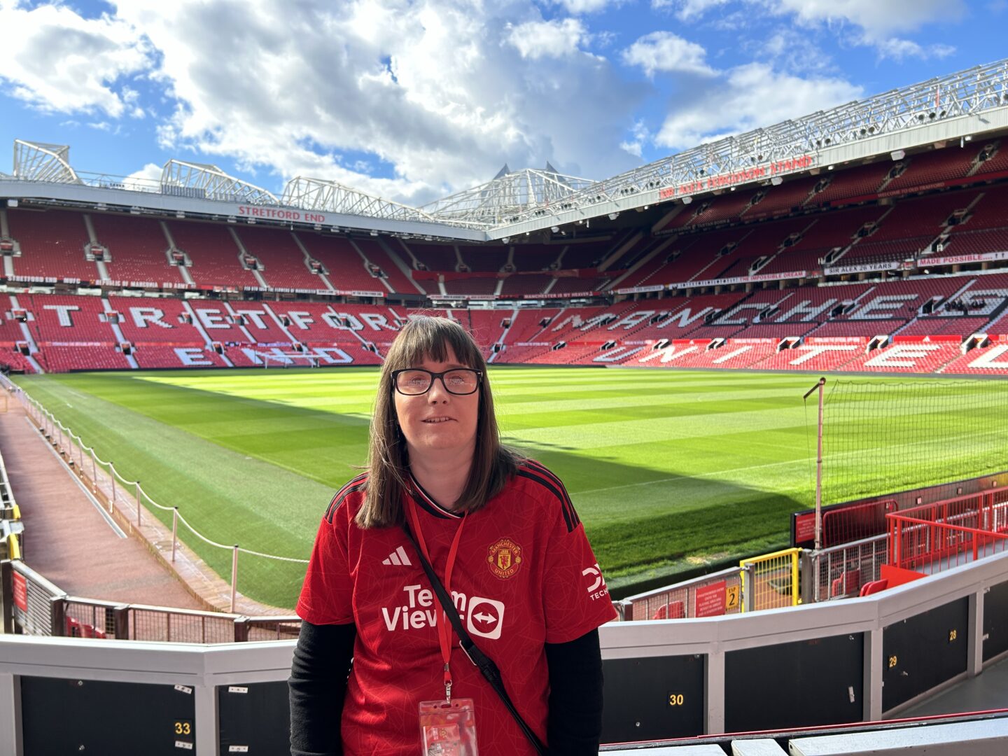 Holly stood in an empty Old Trafford stadium. She’s wearing a red Manchester United shirt and lanyard. The pitch and red seats of the famous Stretford End are visible in the background.
