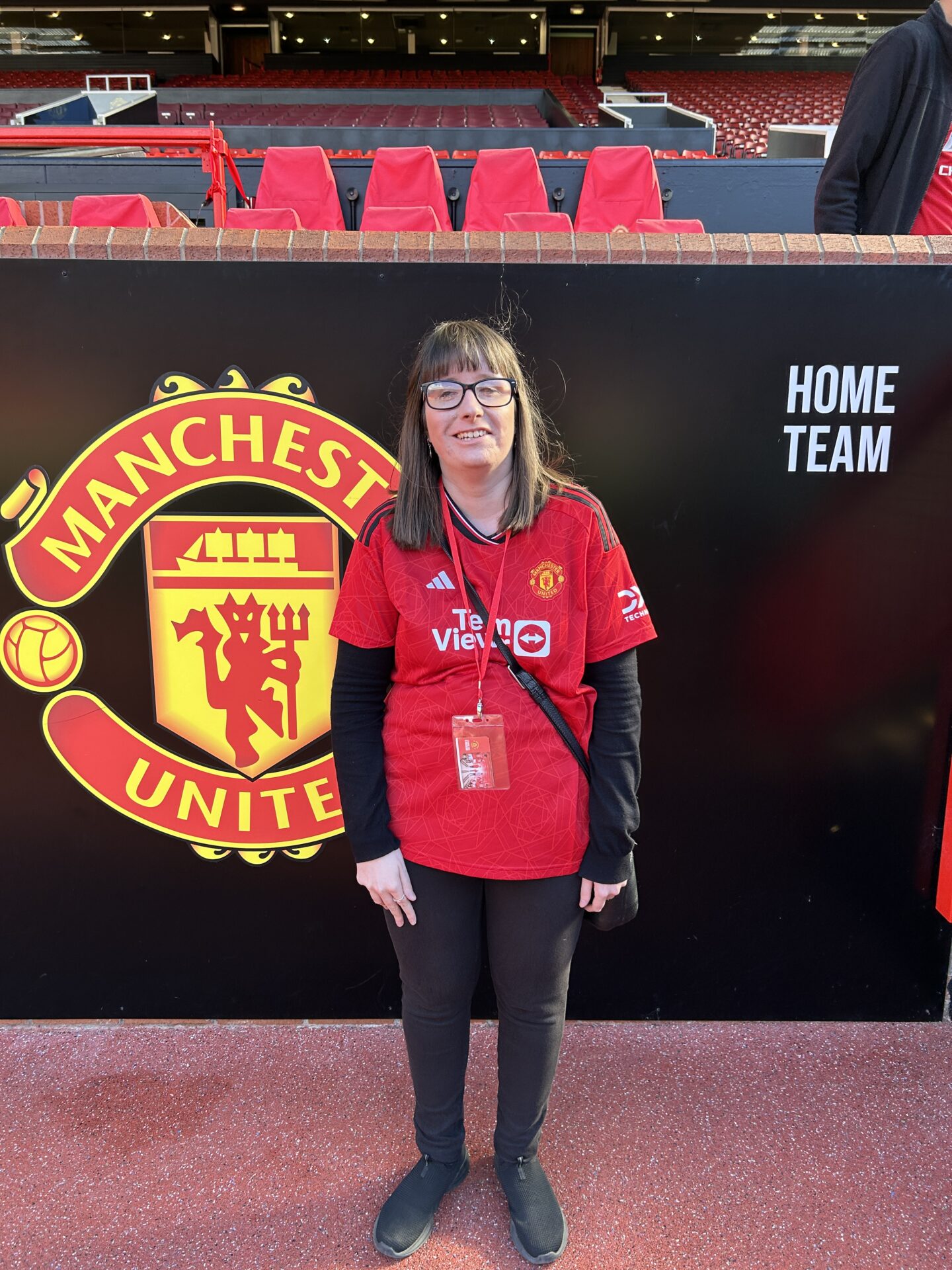 Holly stood in front of the Manchester United sign with the managers dugouts in the background. She has shoulder-length brown hair, wears glasses, a red lanyard and Manchester United shirt, black jeggings and black shoes.