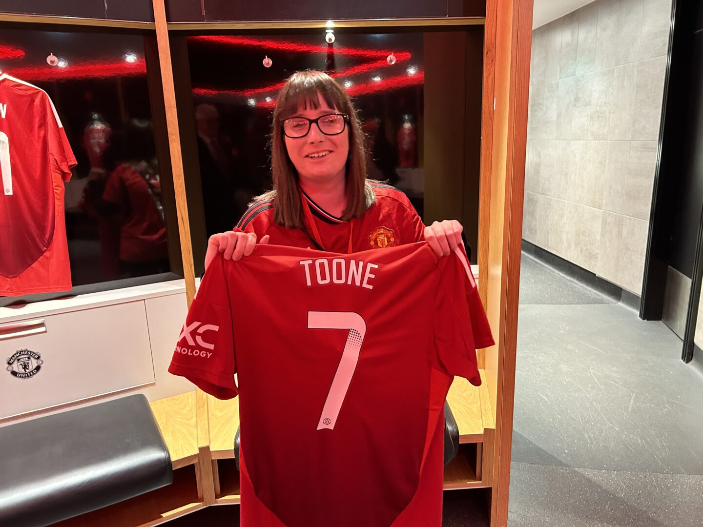 Holly standing in the players changing room at Old Trafford with a big smile on her face. She’s holding up Ella Toone’s shirt. Holly has shoulder-length brown hair, is wearing dark framed glasses and a red Manchester United shirt.