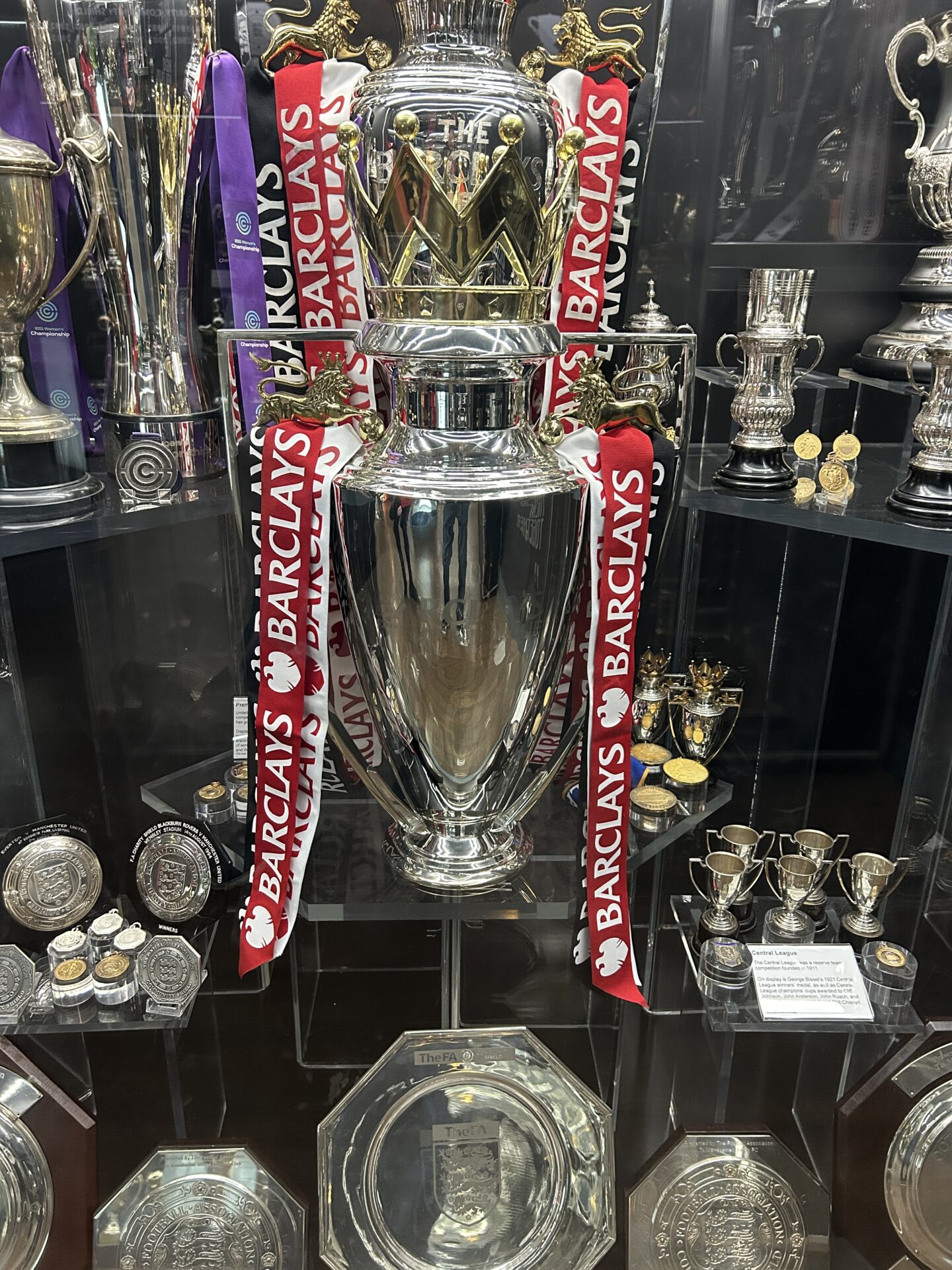 A cabinet filled with various trophies and awards. The centrepiece is the Women’s Premier League trophy, which is a large, silver trophy with a golden crown. It has red and white ribbons on either side with the sponsor Barclays printed on them.