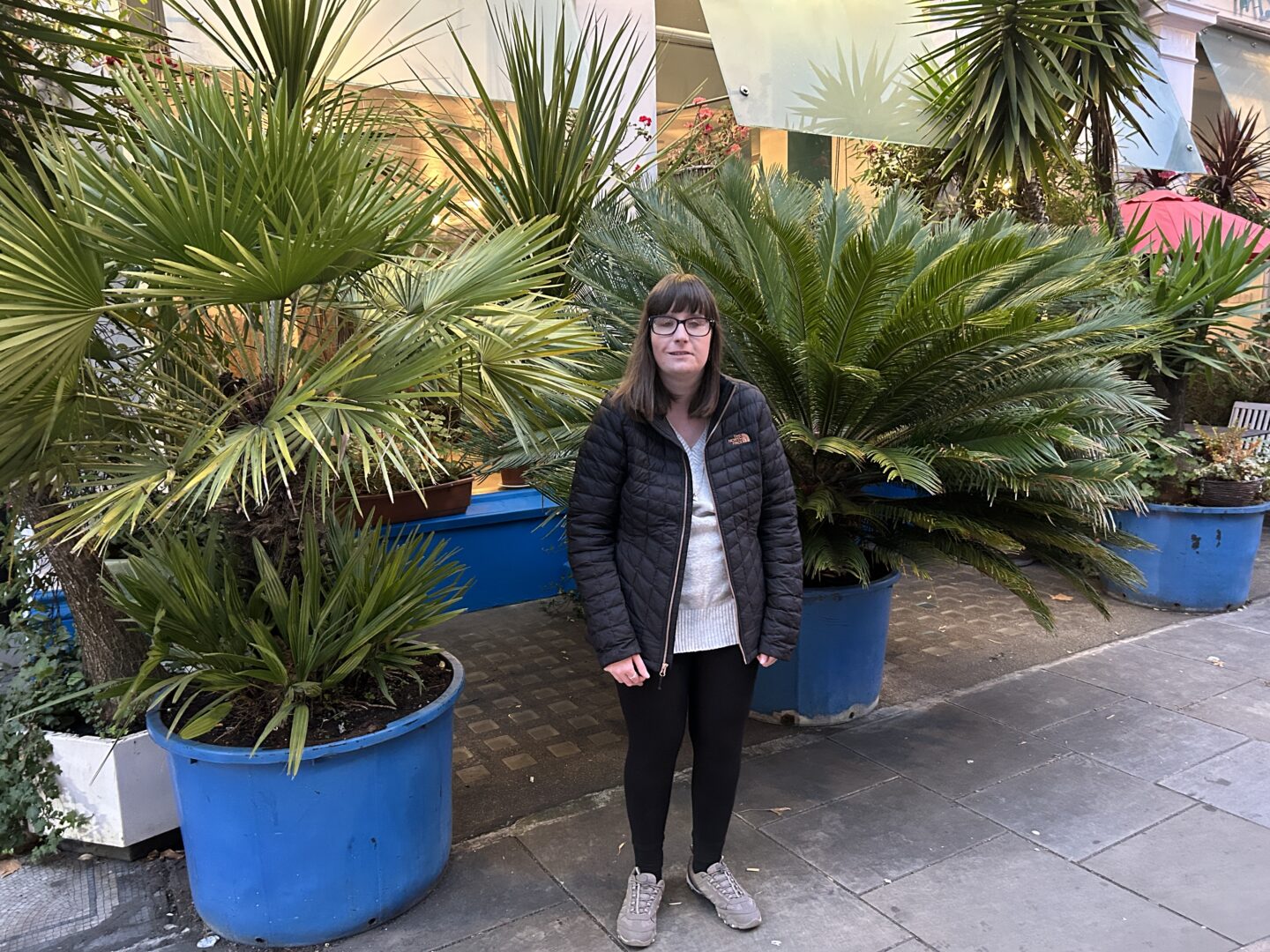 Holly stood in front of green palm trees which are in large blue pots. She has shoulder-length brown hair and wears glasses, a black coat, grey jumper, black leggings and grey trainers.