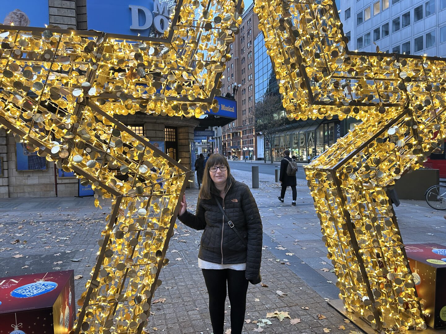 Holly stood in the middle of an ornamental star which is illuminated in gold. She has her hand reaching out towards it and is wearing a black coat and black leggings.