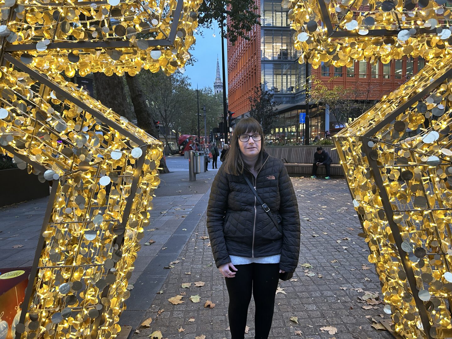 Holly stood in the middle of an ornamental star which is illuminated in gold. She wears a black coat and black leggings.