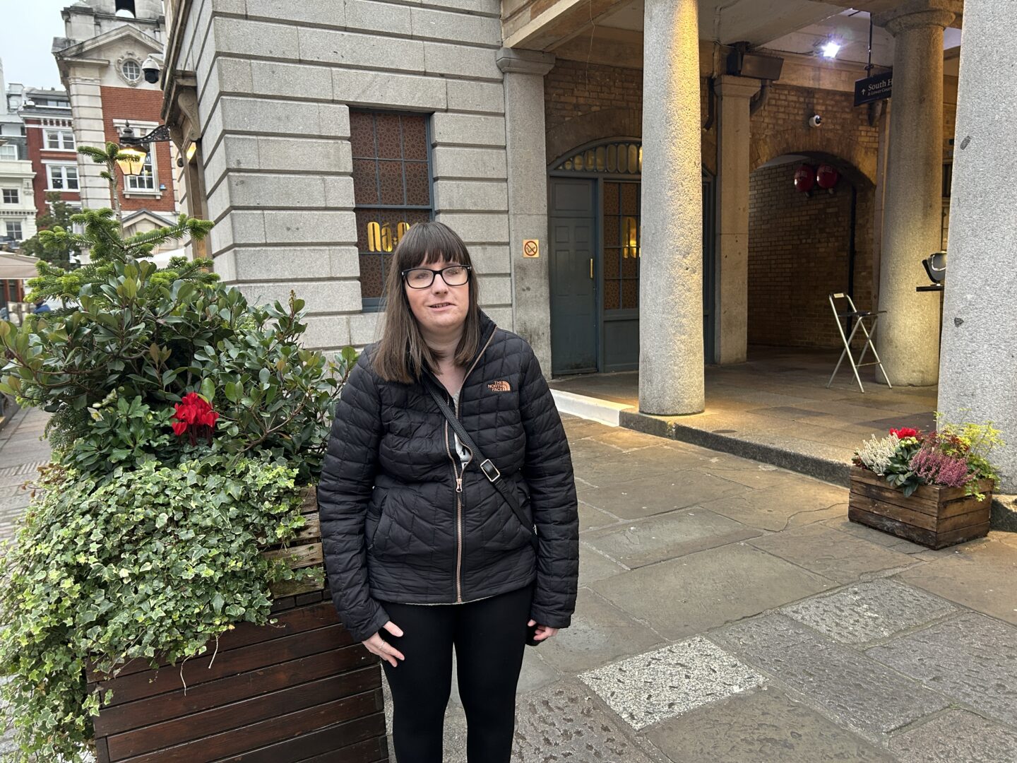 Holly stood next to planters with shrubs inside. She wears a black coat and black leggings.