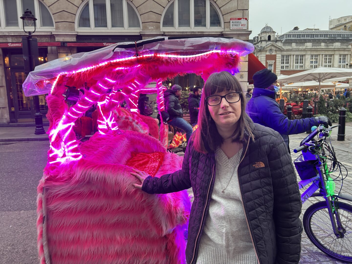 Holly stood with her hand on a rickshaw bicycle which is adorned with pink lights and has fluffy seating. She has shoulder-length brown hair, wears glasses, a black coat and grey jumper and is smiling at the camera.