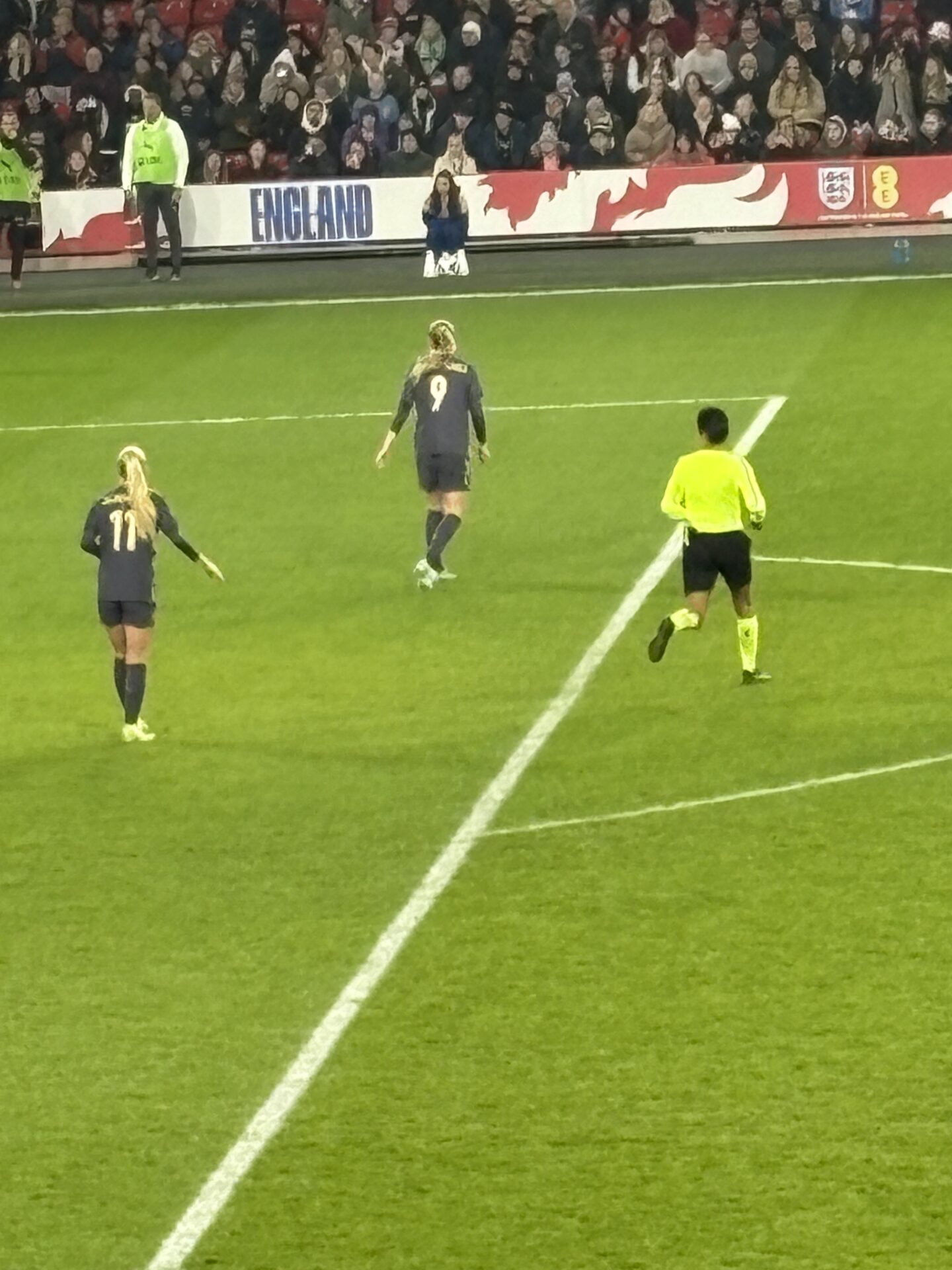Aggie Beever-Jones and Laura Blindkilde Brown, wearing the England purple away kit are running towards the touchline followed by the referee who is wearing a yellow top, black shorts, socks and trainers.