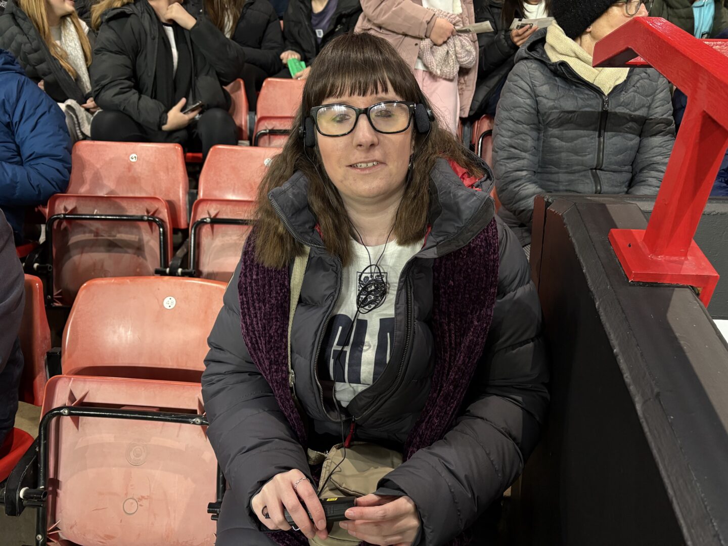 Holly sat in her seat in the stadium, holding her audio commentary device in her right hand. She's wearing a burgundy scarf and a dark blue coat which is partly unzipped to reveal a white England shirt which has the word England in Blue across the front.