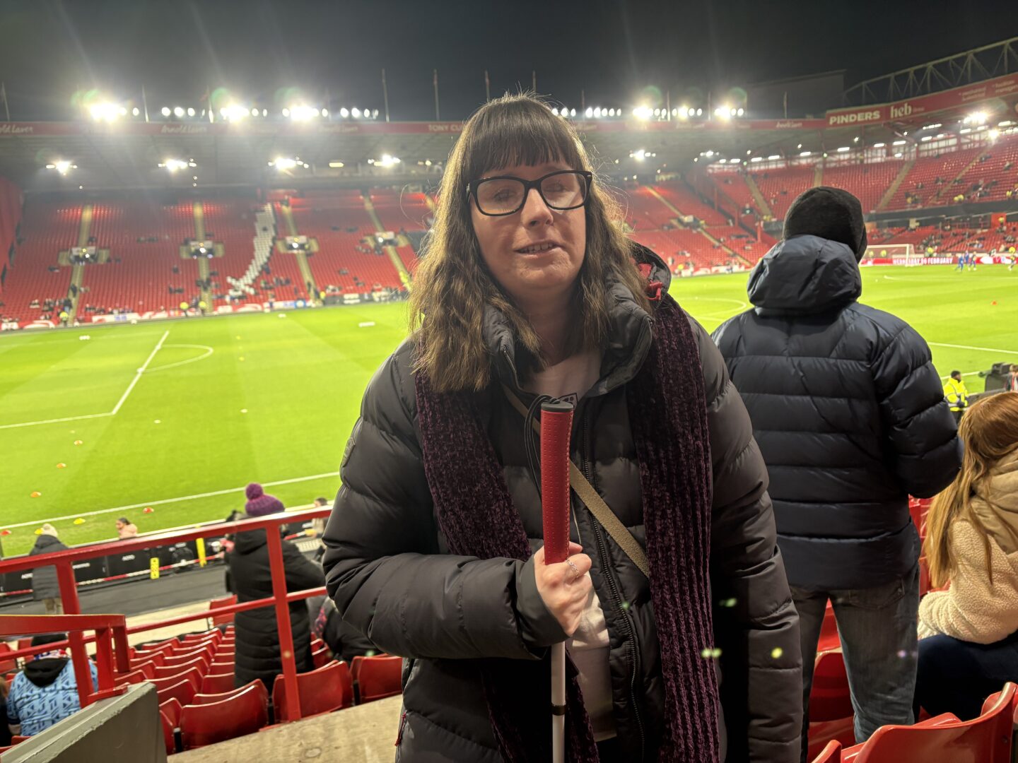 Holly stood in the stadium at Bramall Lane, holding her long cane which has a red handle. The stands and the pitch are visible in the background. She’s wearing a dark blue coat and a burgundy scarf, and her long brown hair is wavy.