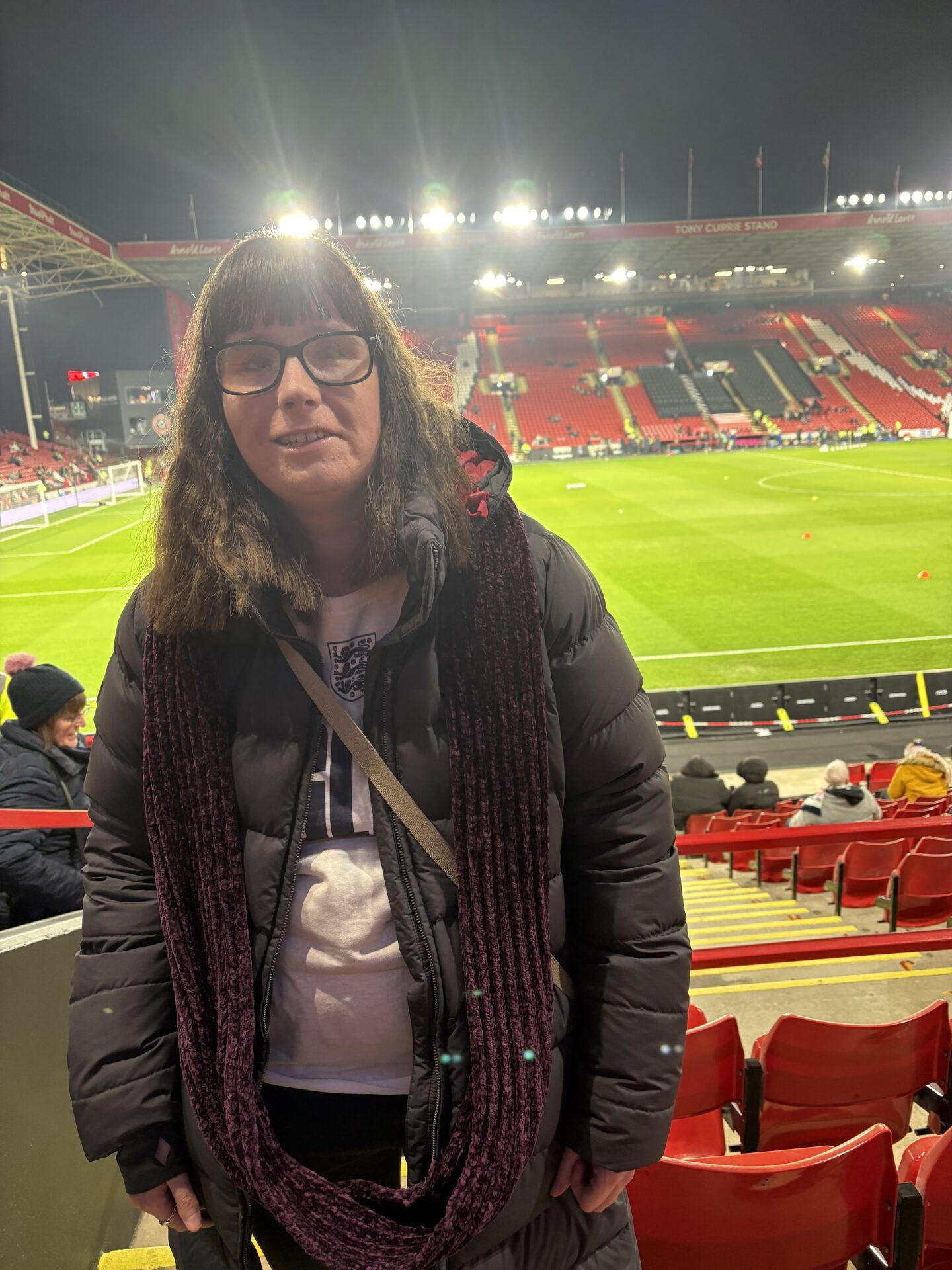Holly stood in the stadium at Bramall Lane. She wears a dark blue coat, white England shirt and a burgundy scarf. Her long brown hair is styled in a wavey do over her shoulders.