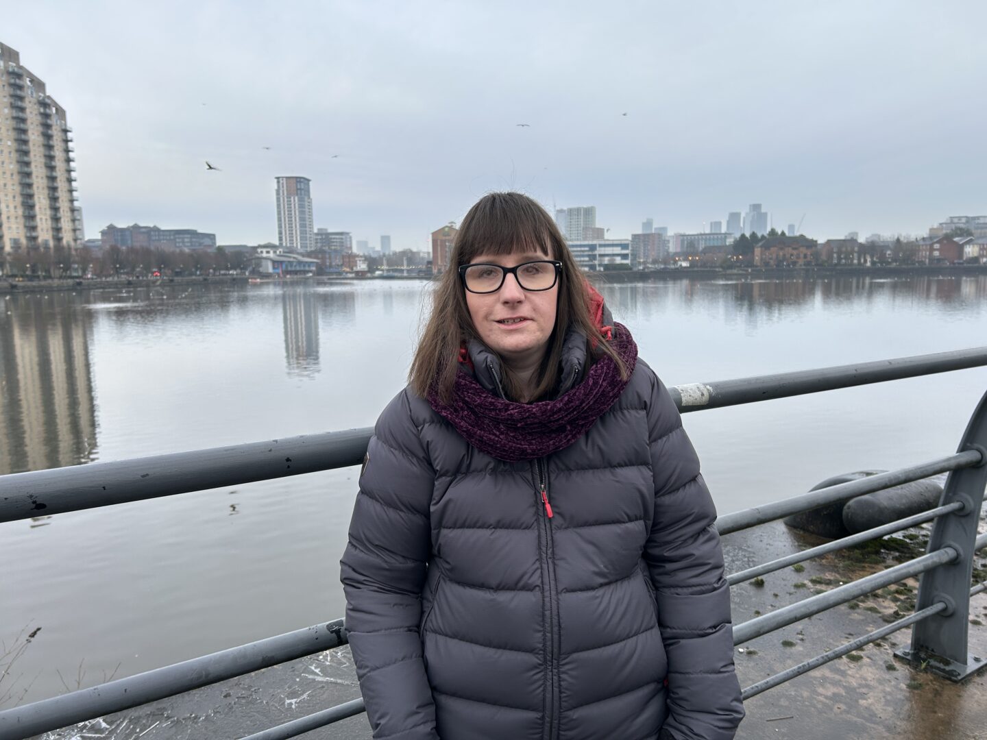 Holly stood on a bridge with tall buildings in the distance. She's wearing a long dark blue coat and a burgundy scarf.