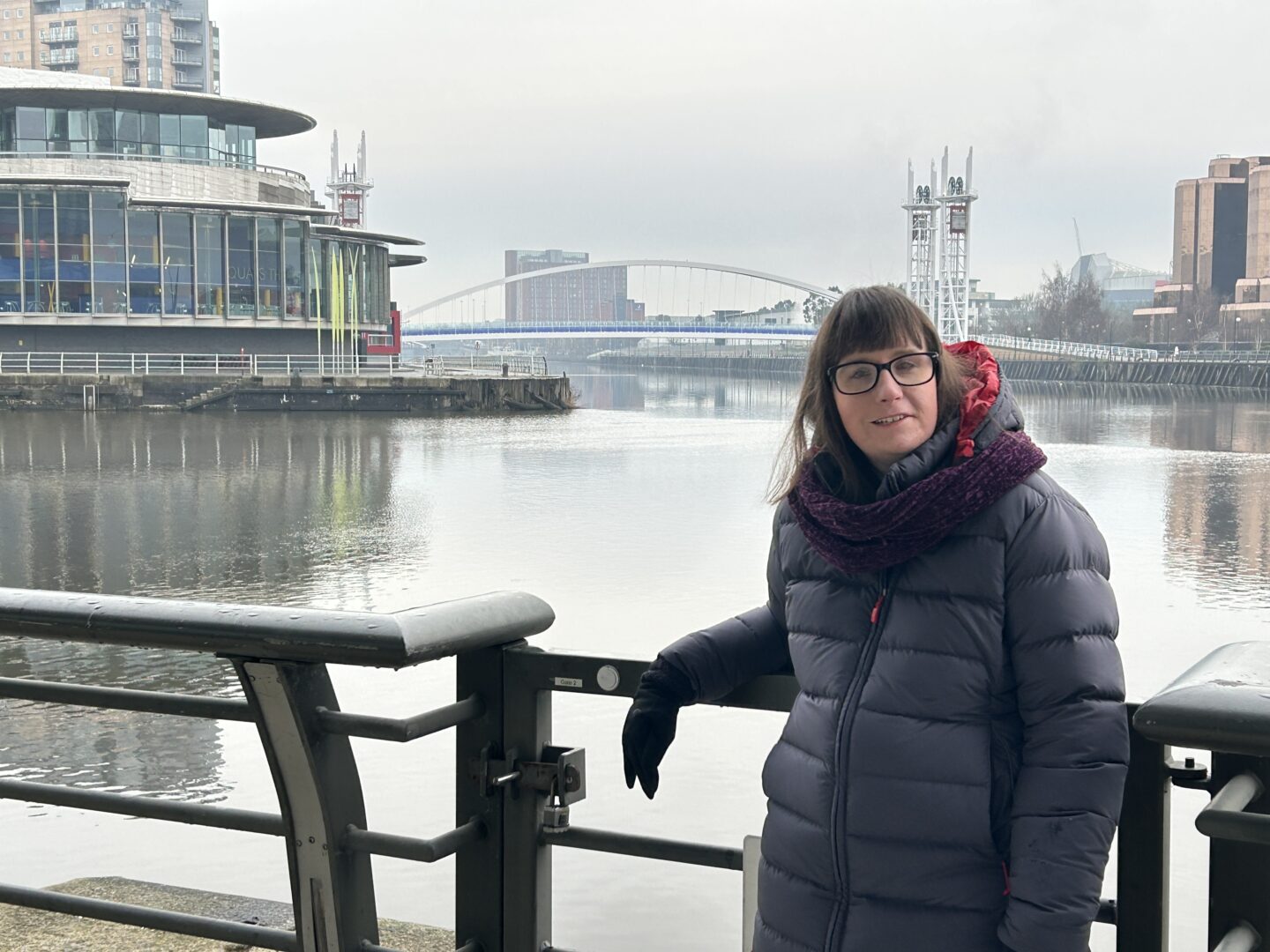 Holly leaning against a railing overlooking a canal with a white arched bridge in the background. Media City can be seen in the distance. She's wearing a long dark blue coat, burgundy scarf and black gloves.
