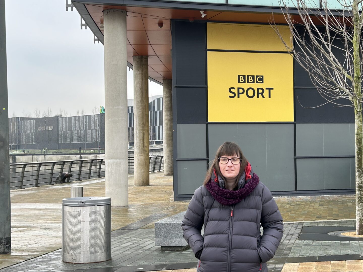 Holly, who wears a dark blue coat and a burgundy scarf stands with her hands in her pockets in front of a yellow BBC sport sign. A canal and buildings can be seen in the distance.