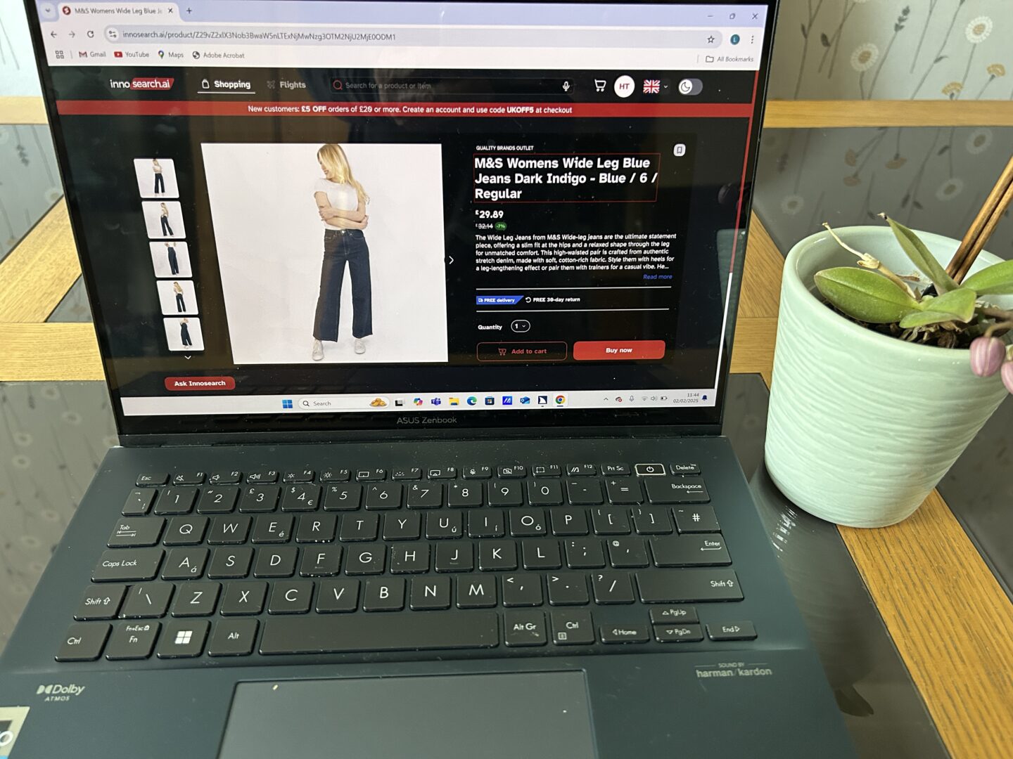 A product page for an item on Innosearch displayed on a laptop screen. The product is some M&S Women’s Wide Leg Blue Jeans. The page shows a large image of a model wearing the jeans, with smaller photos surrounding it, along with relevant info such as the price and the product description. There is an orchid next to the laptop.