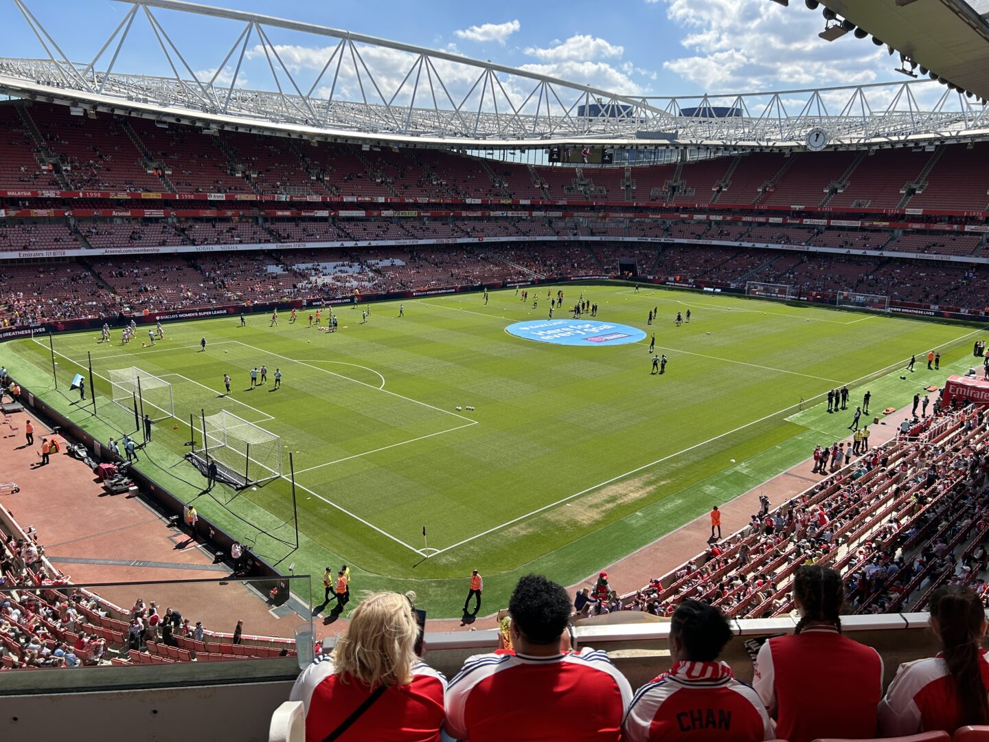 A view looking down on the pitch at Emirates Stadium. The stands are partially filled with fans. Players can be seen warming up on the pitch, and there is a large circular blue and white emblem in the centre of the pitch.