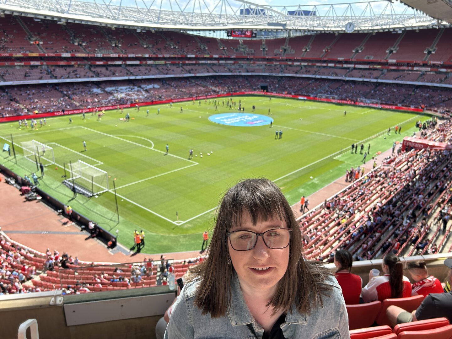 Holly stood in the Emirates Stadium with a view of the pitch behind her. She’s wearing a light blue denim jacket and is smiling at the camera.