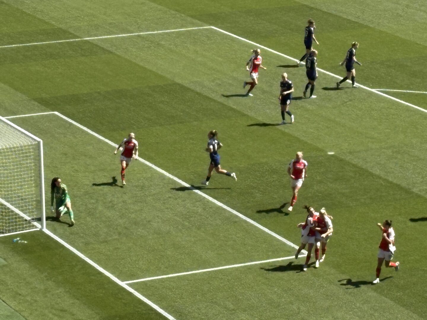 Two Arsenal players hugging each other after celebrating a goal. Other Arsenal players can be seen running towards them to join in the celebrations, whilst Manchester United players are walking away. The Arsenal players are wearing their red home shirts, and the Manchester United players are wearing their blue away shirts.