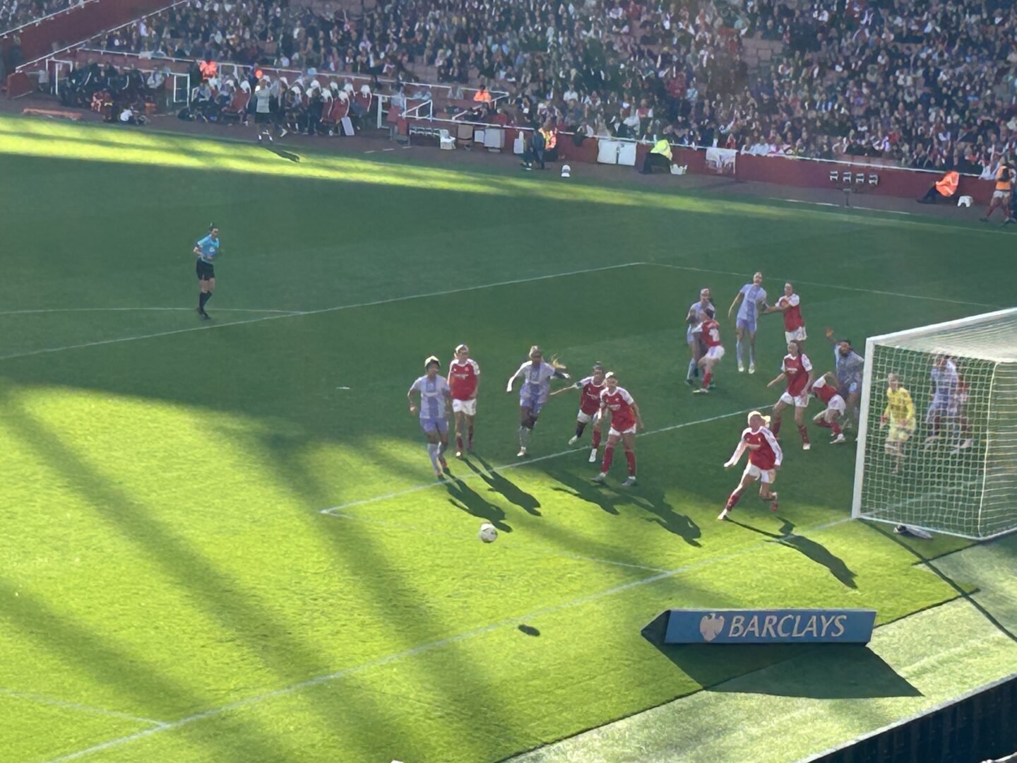 Arsenal and Brighton players near one of the goals on the pitch. The referee can be seen watching from a distance. There is a Barclays advertising board near the corner of the pitch. Fans fill the stands, watching the action.