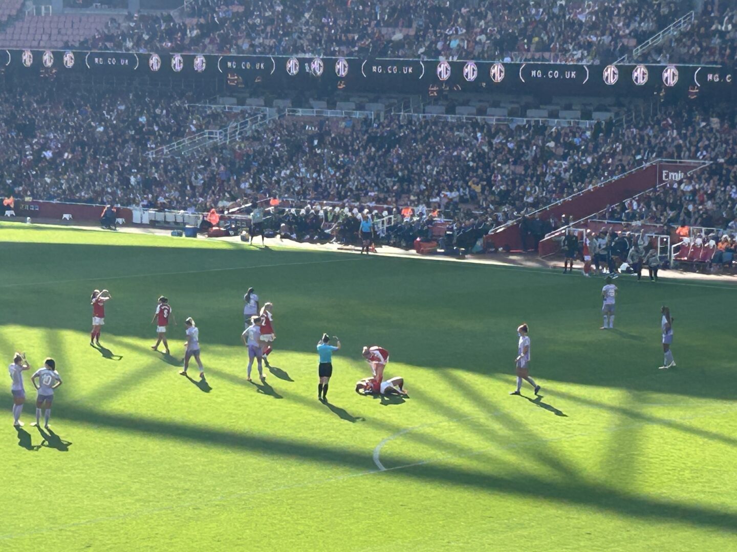Arsenal and Brighton players on the pitch at Emirates Stadium. Arsenal are in their traditional red and white home kit, whilst Brighton are in their purple away kit. Fans can be seen enjoying the match around the stadium on a sunny day.