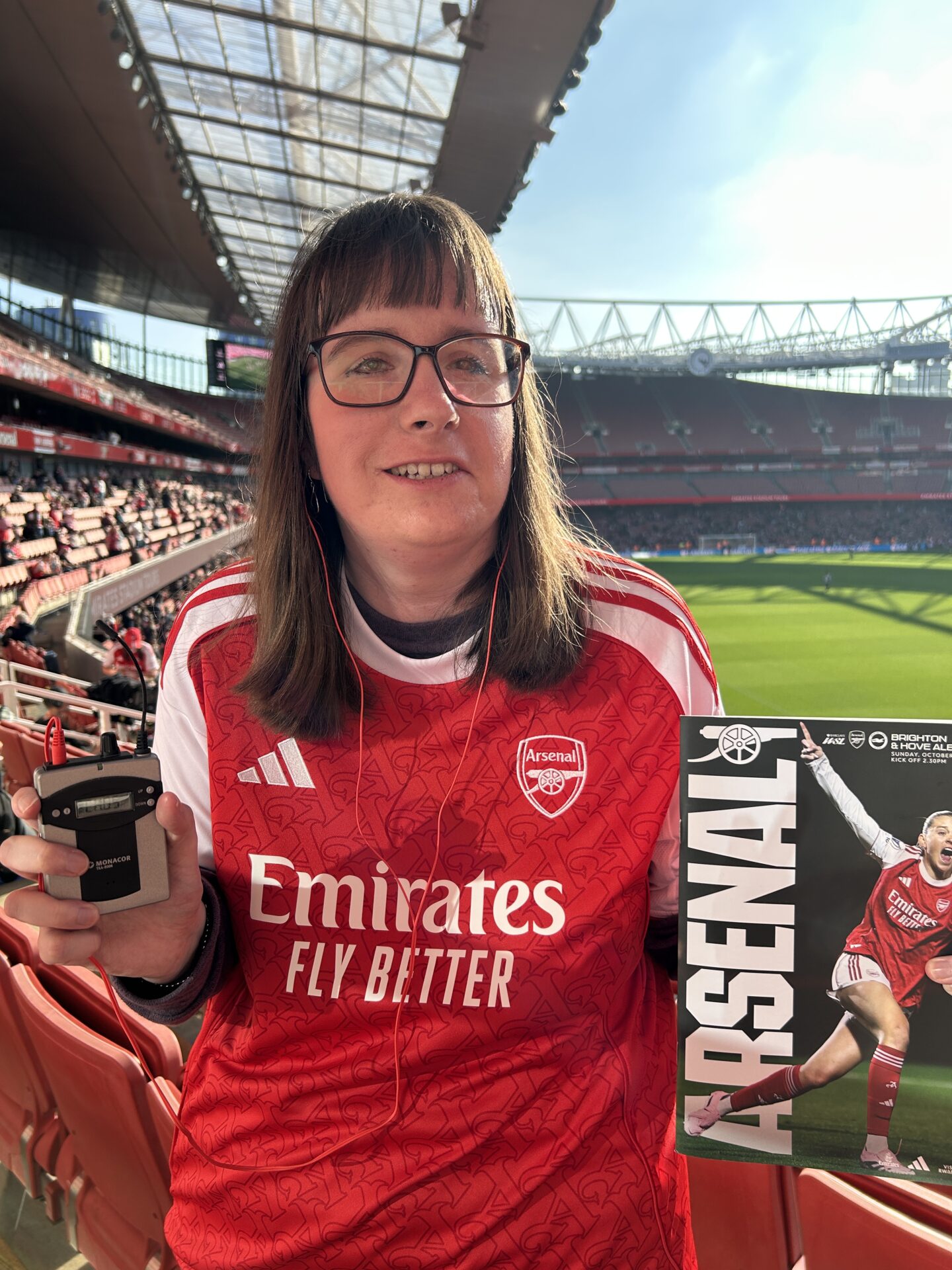 Holly standing in the stands at Emirates Stadium with the pitch and stadium seats behind her. She’s wearing a red and white Arsenal shirt and light blue jeans. She’s holding the Arsenal matchday programme and audio descriptive commentary receiver, and is smiling at the camera.