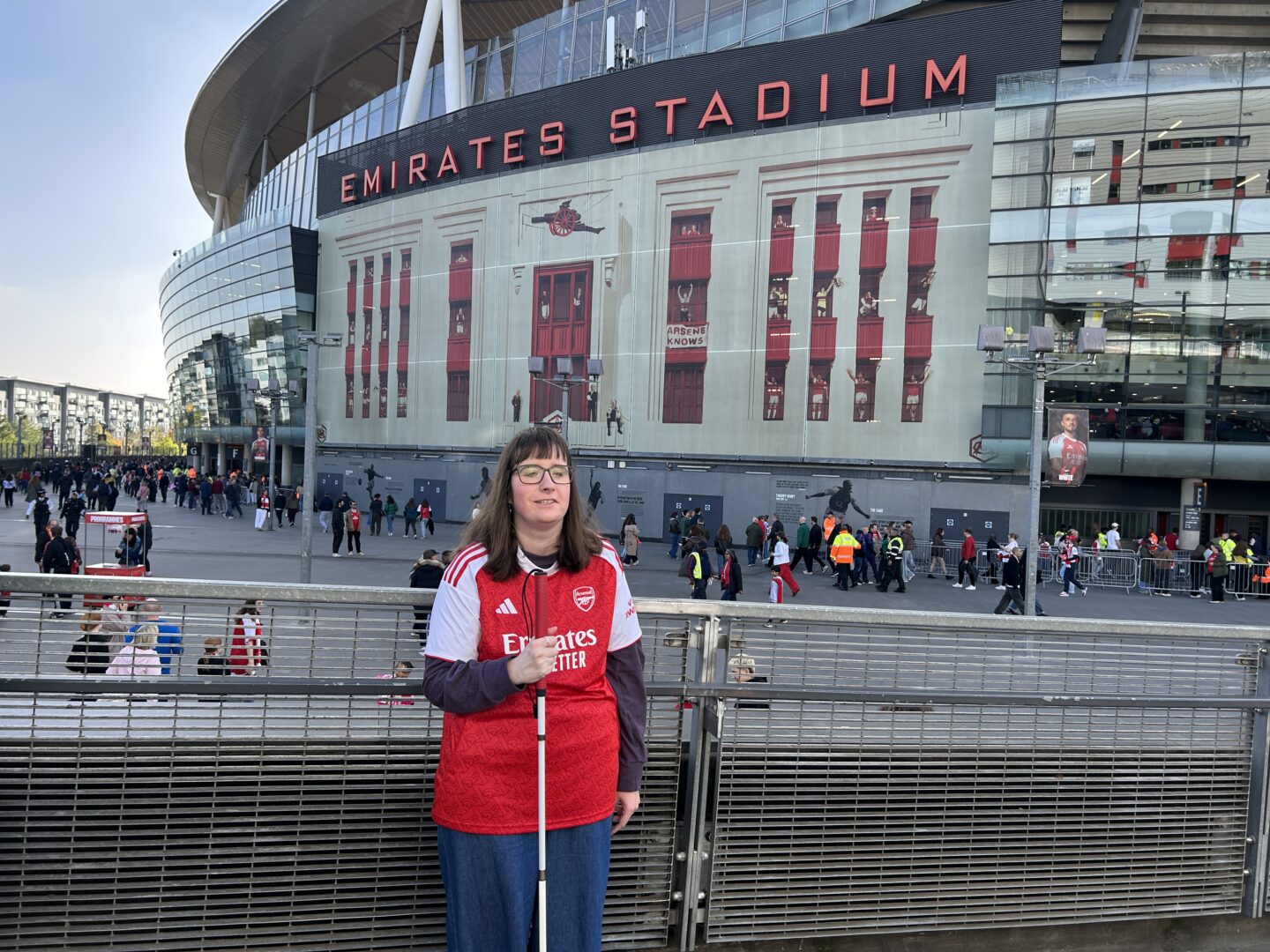 Holly stood outside Emirates Stadium. She wears glasses, a red and white Arsenal shirt which features the Arsenal logo on the left and the sponsor “Emirates” across the front, a purple top, and light blue jeans. She’s holding her white cane in her right hand and is smiling at the camera.