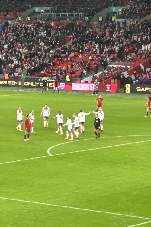 England and China players on the pitch. England are wearing white shirts and blue shorts, and China wear red kits. They are gathered near the penalty box area, with England players celebrating a goal. A referee dressed in black stands nearby with some players, while fans fill the stands in the background.