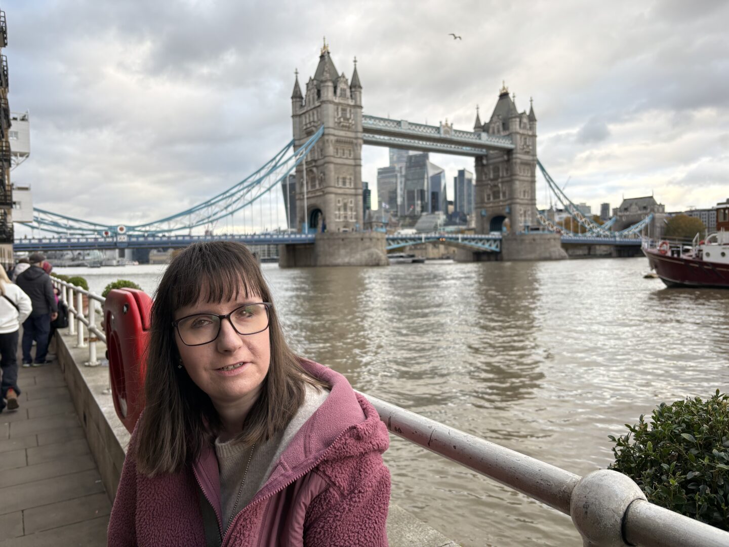 Holly sat on a wall with a railing and the River Thames behind her. Tower Bridge is in the distance, and a boat can be seen coming into view. She’s wearing a pink fleece jacket and a beige jumper.