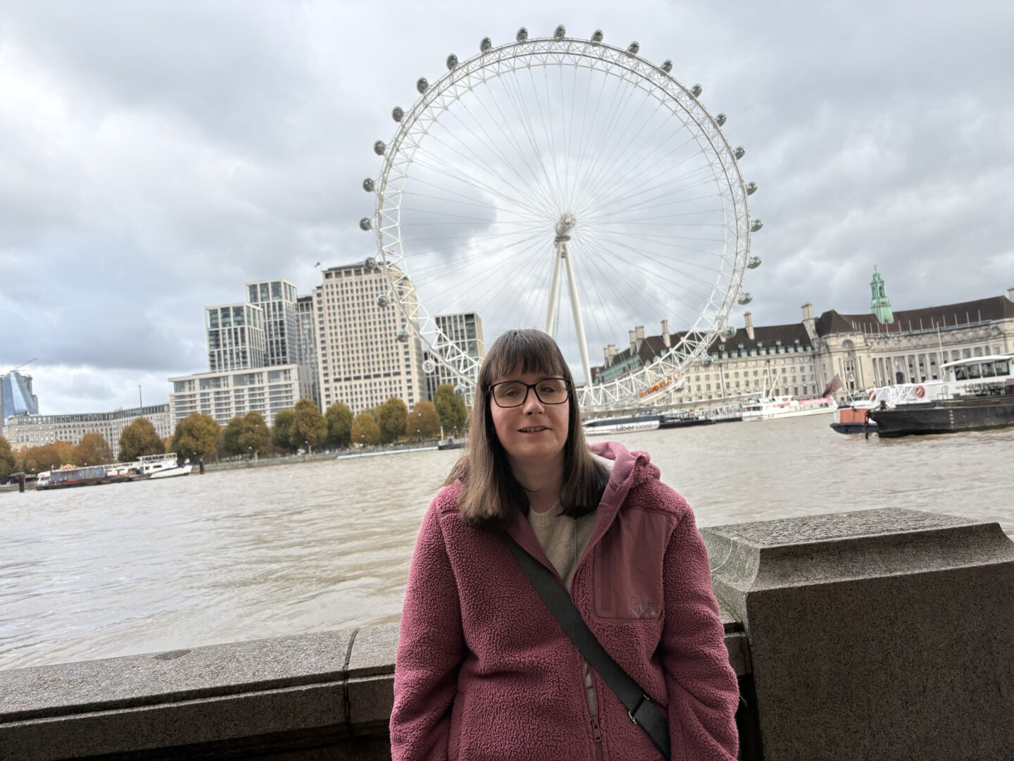 Holly stood in front of a stone wall with the London Eye in the background. She’s wearing glasses and a pink fleece jacket with a handbag across her shoulder. Canal boats are on the River Thames behind her with buildings in the distance.