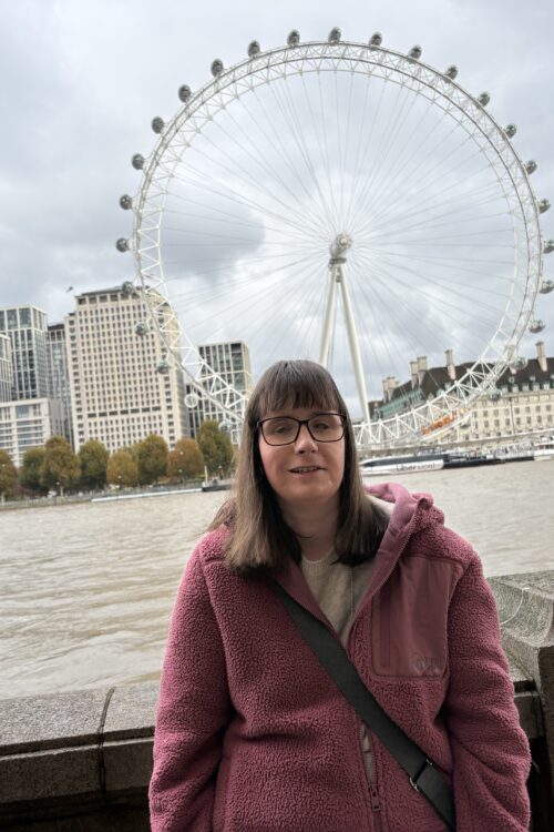 Holly stood in front of a stone wall with the London Eye in the background. She’s wearing glasses and a pink fleece jacket with a handbag across her shoulder. Canal boats are on the River Thames behind her with buildings in the distance.