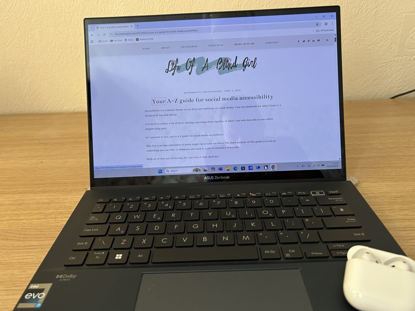 A laptop placed on a wooden desk. The laptop screen displays Holly’s blog post titled ‘Your a-Z guide for social media accessibility’. On the bottom right hand corner of the laptop, there is a white case with Apple AirPods inside.