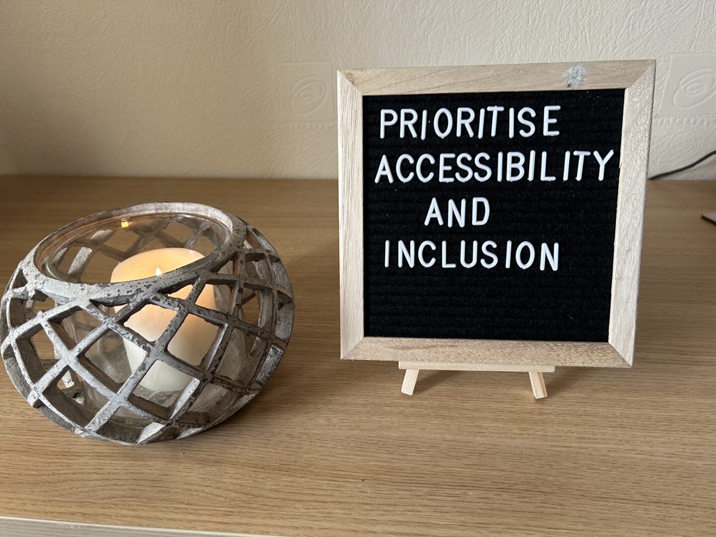 A candleholder with a lit white candle is placed on a desk next to a felt letterboard on a small wooden easel. The board has white letters that spell out ‘prioritise accessibility and inclusion’.