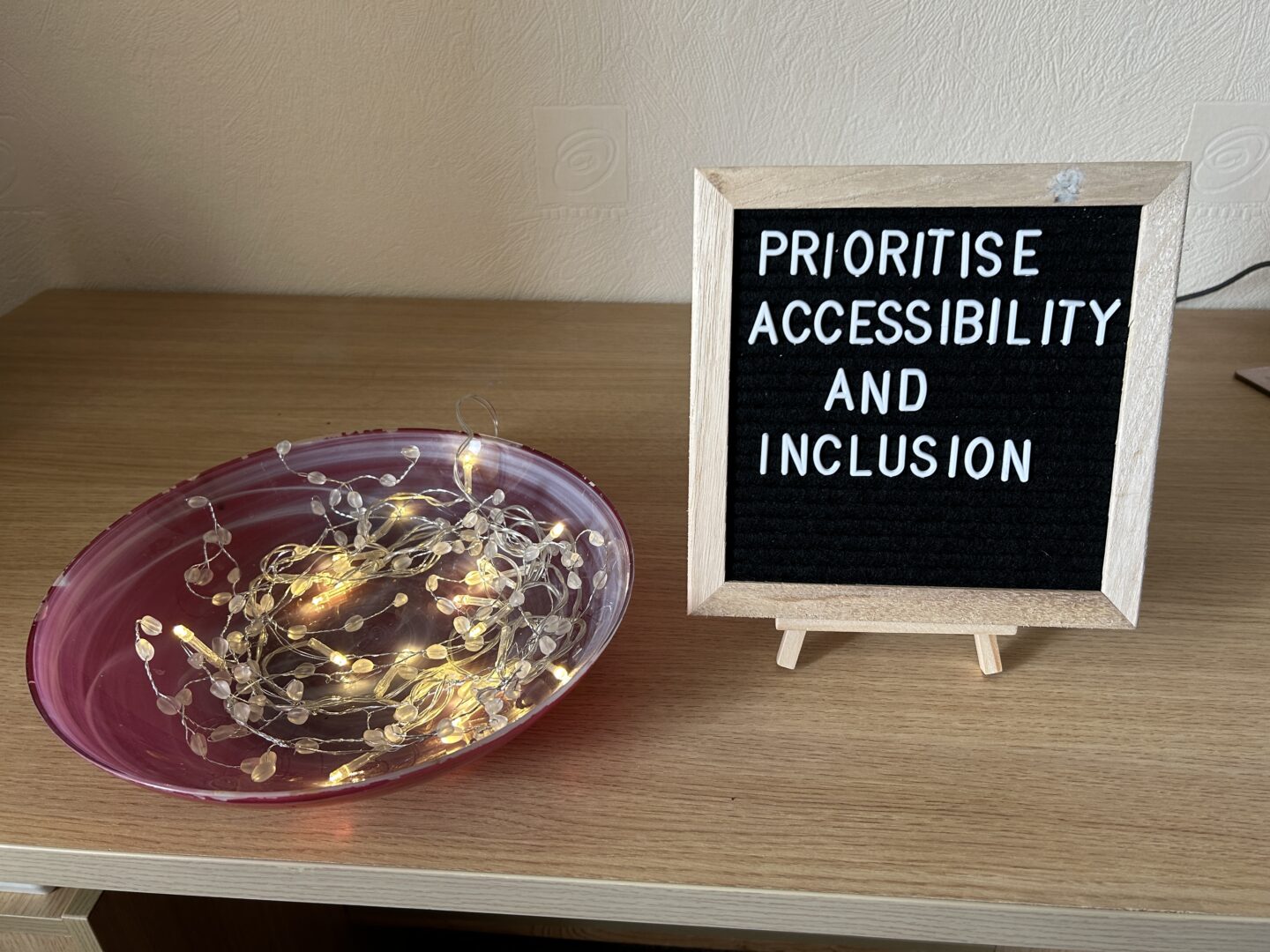 A small black letterboard with a light wooden frame on a mini easel sits on a desk. The board has white letters which spell out ‘prioritise accessibility and inclusion’. There is a round, pink and white swirled bowl filled with a string of small fairy lights that are switched on next to it.