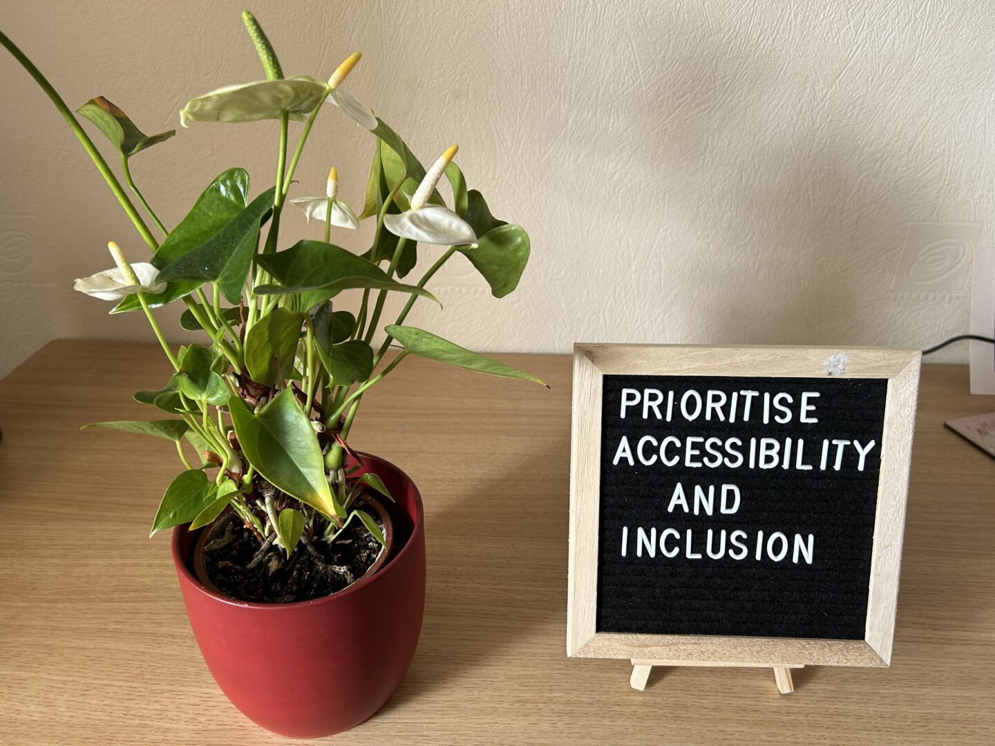 A black felt letterboard in a light wooden frame on a mini easel sits on a desk with a leafy plant with white flowers in a red pot next to it. The board has white letters that spell out ‘prioritise accessibility and inclusion’.