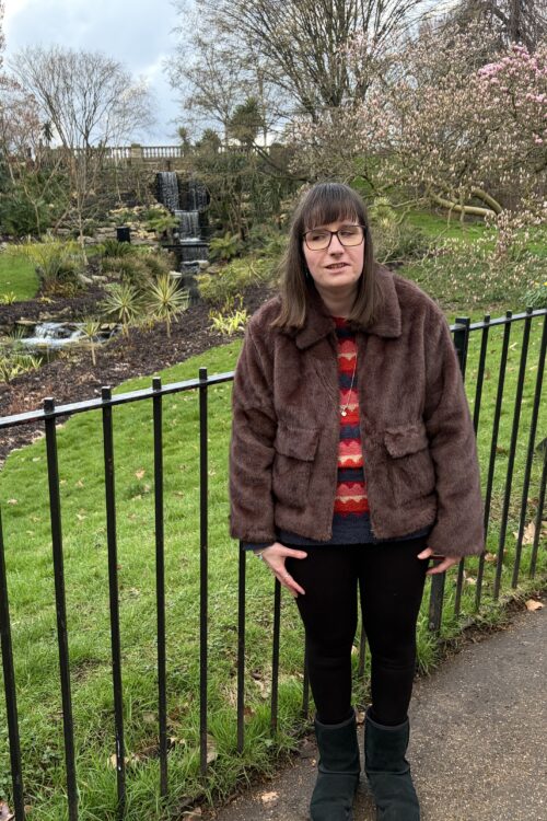 Holly stood in front of black railings. There is a beautiful garden of palm trees, luscious green grass, trees in blossom and a beautiful waterfall behind her. She wears a brown faux fur jacket, a red and navy patterned jumper, black leggings and black boots, and is smiling at the camera.