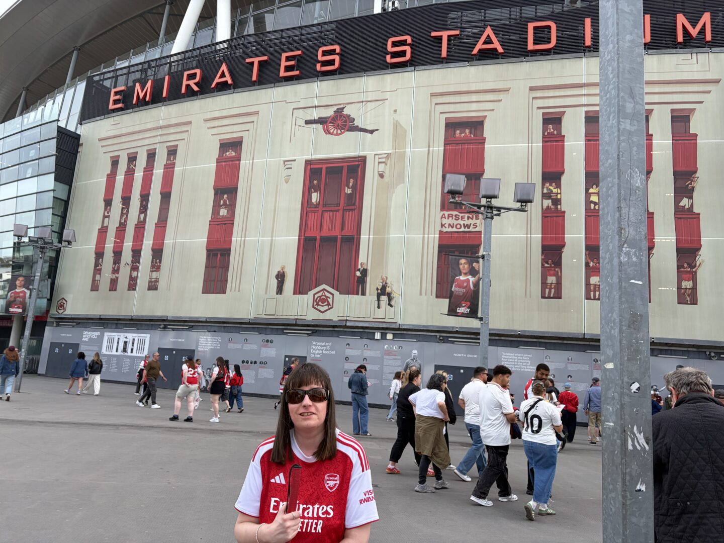 Holly stood outside Emirates Stadium. She wears sunglasses, a red and white Arsenal shirt which features the Arsenal logo and the sponsor 'Emirates' across the front, black leggings and black trainers. She’s holding her long cane and is smiling at the camera. The stadium is behind her, and large red letters across the top spell 'Emirates Stadium.'