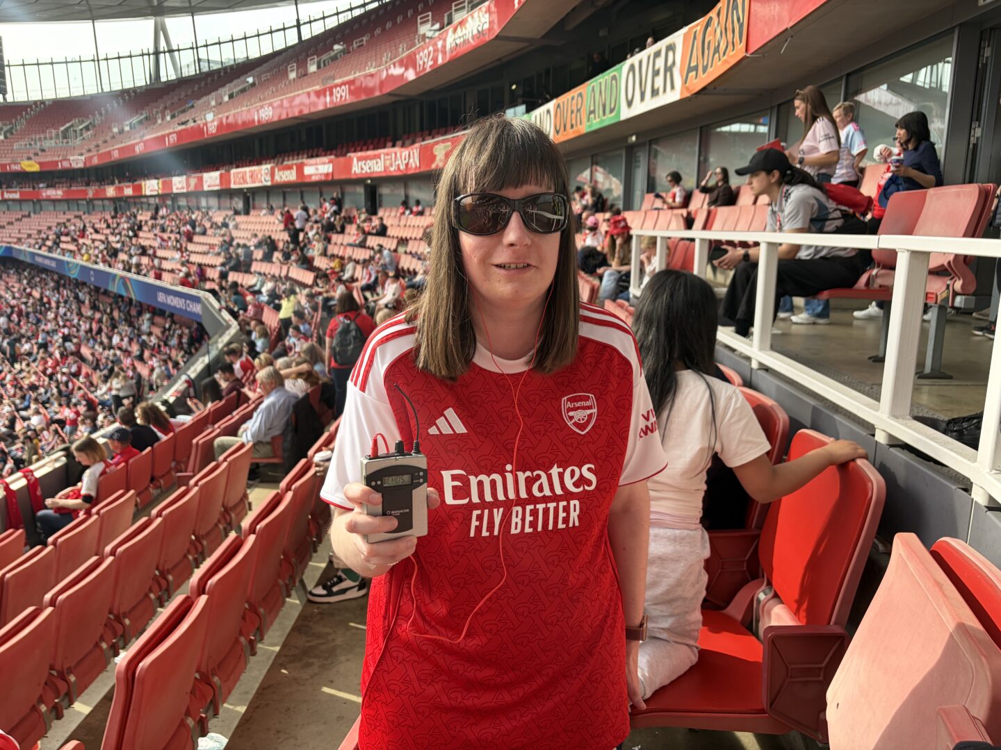 Holly stood in the stands at Emirates Stadium. She’s holding her audio descriptive commentary receiver and is smiling at the camera. Rows of red seats can be seen filling the stands in the background behind her and banners adorn the stadium. She wears sunglasses, a red and white Arsenal shirt and black leggings.