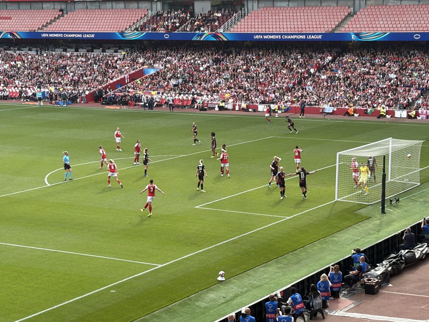 Arsenal and Lyon players on the pitch at Emirates Stadium. Arsenal players who are in their red and white home kit are walking away after scoring a goal, whilst Lyon players are in a black kit. The ball is in the back of the net, with the Lyon goalkeeper in a yellow kit looking towards the ball with a look of “I should have saved that!”. The referee is positioned to the left side of the penalty area. Fans can be seen enjoying the match from the stands. A banner reads 'UEFA Women’s Champions League.'