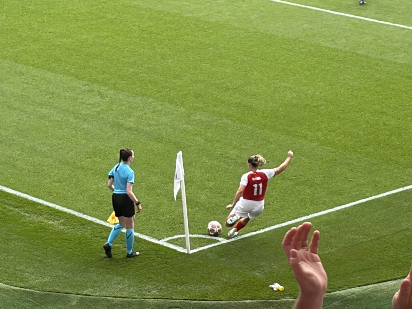 Arsenal’s Katie McCabe taking a corner kick. Her blond hair is tied back in a ponytail, and she wears a red and white jersey with the number 11 visible on her back, white shorts and red socks. The linesperson is stood to her left side.