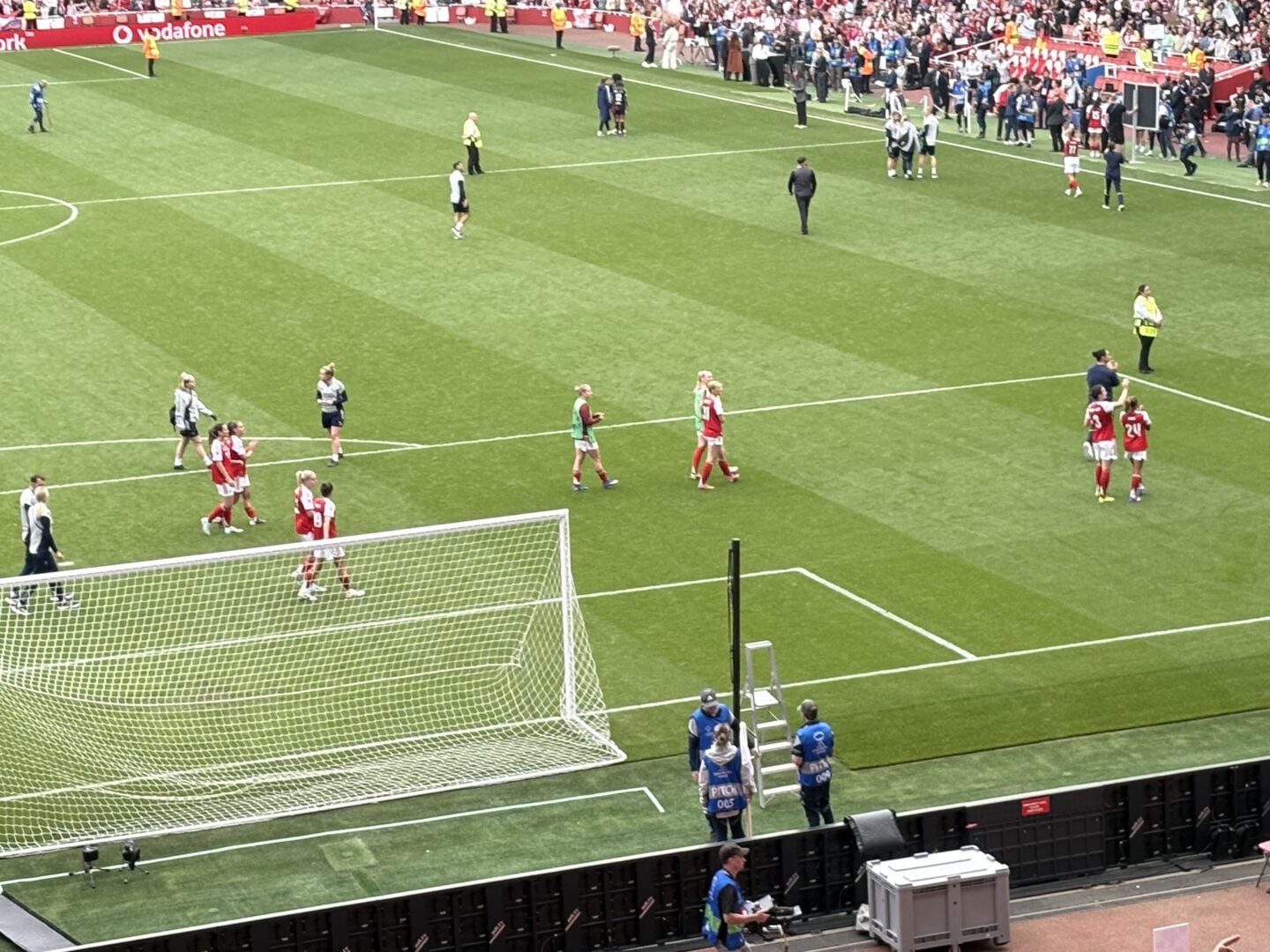 Arsenal players on the pitch clapping and waving, showing their support to the fans at the end of the match. Some of the rest of the squad have also joined them on the pitch. Fans can be seen celebrating in the stands in the background.