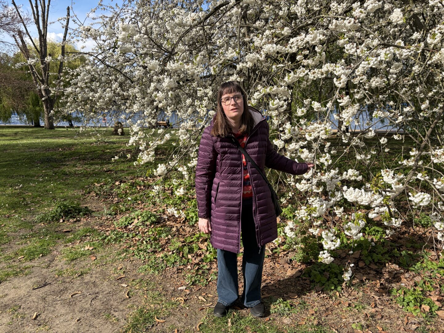 Holly stood in front of a white cherry blossom tree with one hand stretched out holding a branch. She wears a burgundy coat, a multicoloured patterned jumper, dark blue jeans and black trainers.