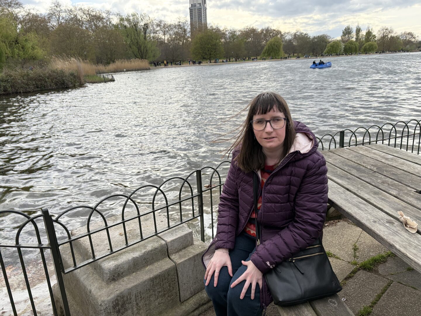 Holly sat on a bench with a lake behind her and an iron fence surrounding it. She wears a burgundy coat, a multicoloured patterned jumper, dark blue jeans and a black crossbody bag. There is a small pedal boat on the lake, and a tree-lined path in the distance.