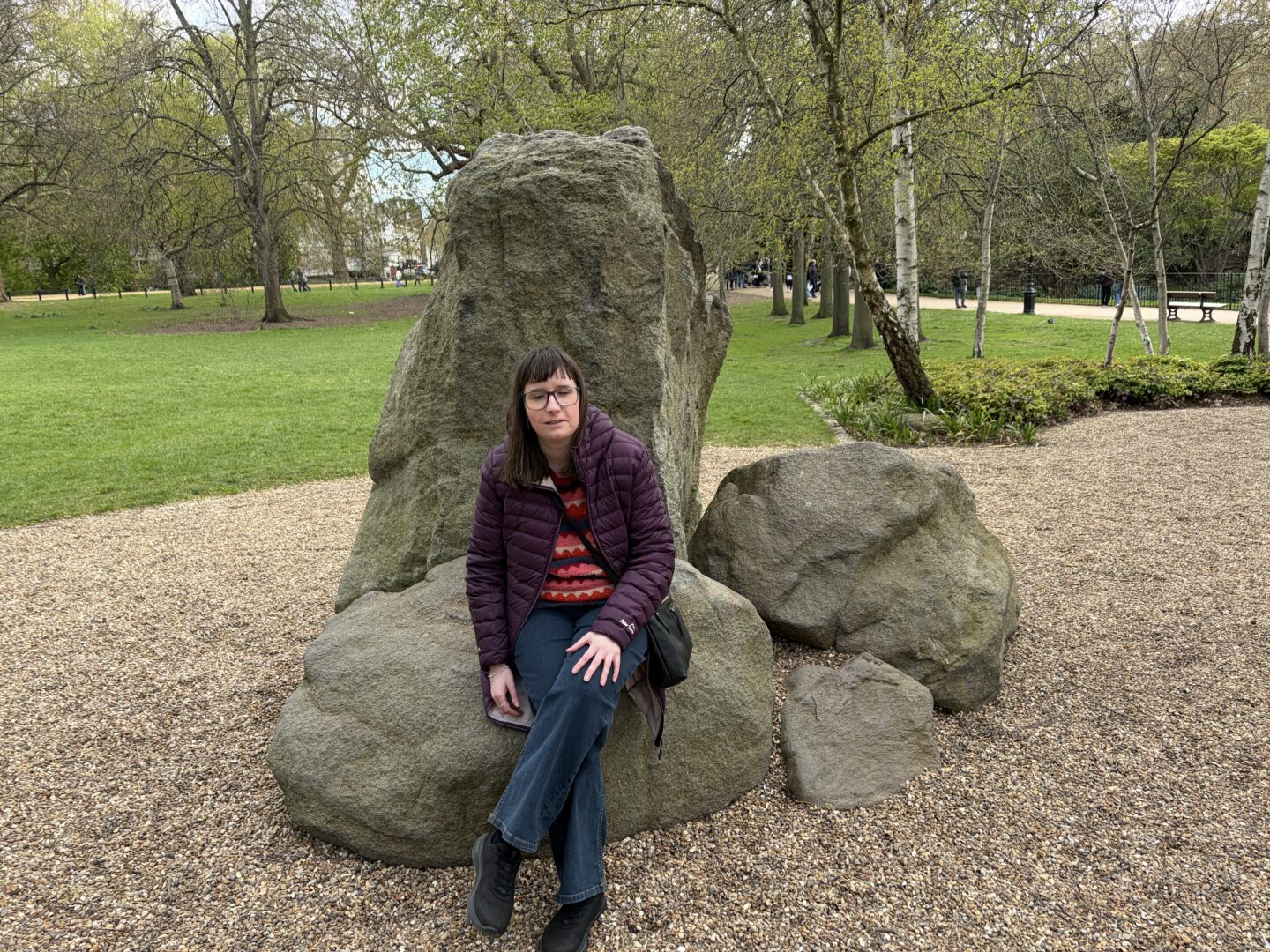 Holly sat on a large rock with smaller rocks to the side. The ground is covered in small brown gravel around the rocks. She wears a burgundy coat, a multicoloured patterned jumper, dark blue jeans, black trainers and a black crossbody bag. She has one leg crossed over the other. There are trees scattered in the distance.