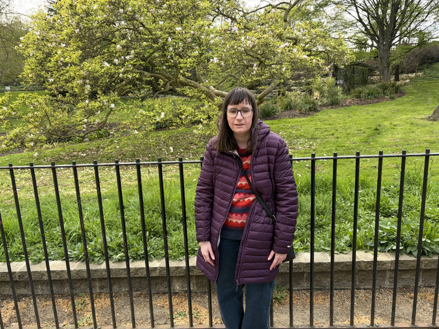 Holly stood in front of a black metal fence with a white cherry blossom tree and a green garden filled with shrubbery in the background. She wears a burgundy coat, a multicoloured patterned jumper, dark blue jeans and a black crossbody bag.