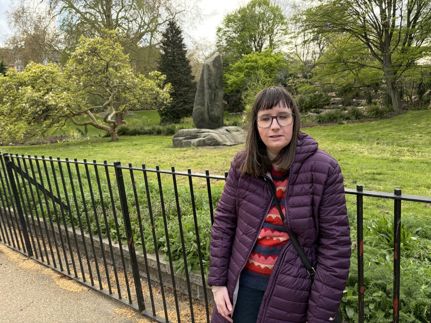 Holly stood in front of a fence in a park. She has shoulder-length brown hair, wears glasses, a burgundy coat, a multicoloured patterned jumper, dark blue jeans and a black crossbody bag. There are lots of trees, spring foliage and a cluster of rocks behind her.