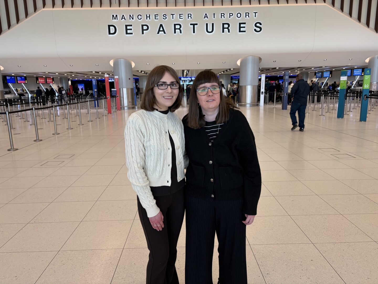 Holly and Elin standing in the terminal at Manchester Airport. Elin wears glasses, a cream cardigan, a black top and black trousers. Holly wears glasses, a black cardigan, a black top with white stripes, and black trousers. A sign is high on the wall behind them, that reads “Manchester Airport departures.”