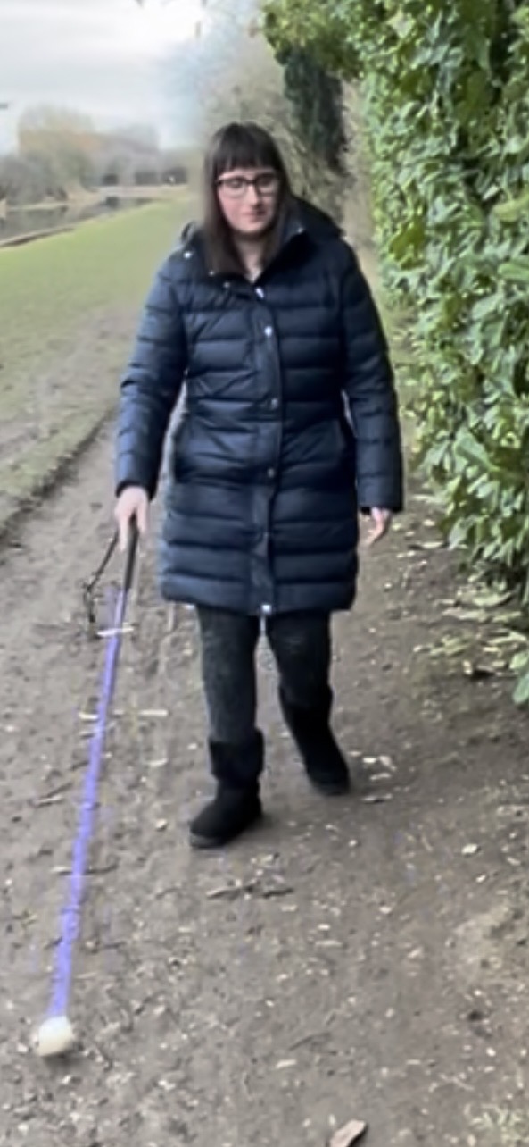 Holly walking along a path, using her purple cane. She’s wearing black-framed glasses, a long dark navy puffer coat, dark trousers and black boots. A hedge runs alongside the path beside her, and there is a grassy bank and a canal with trees and bushes in the distance.