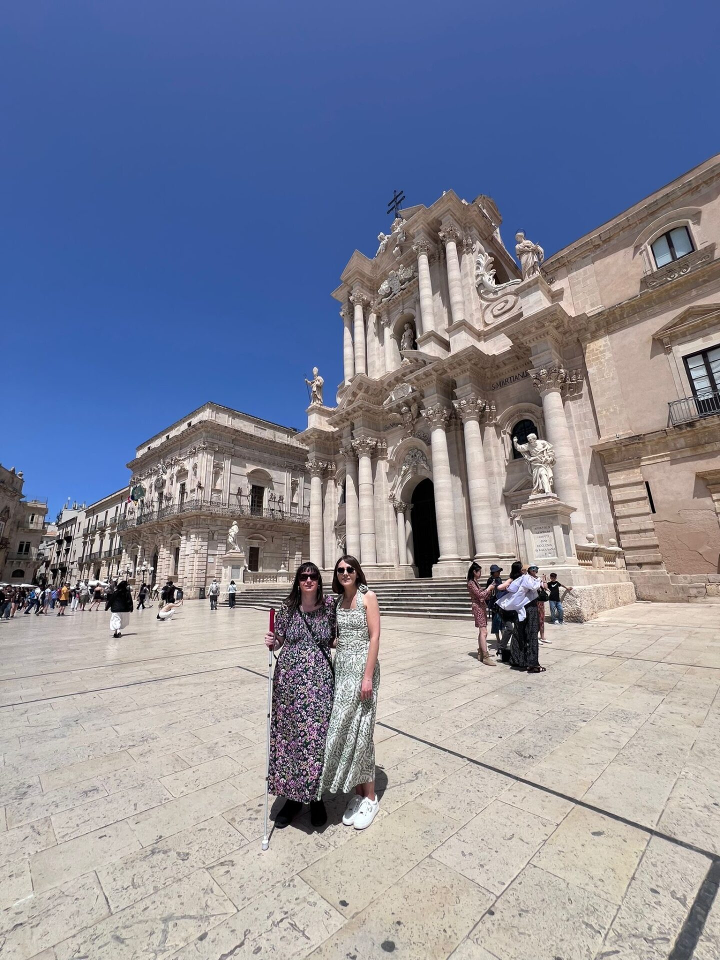 Holly and Elin standing with their arms around each other in a large sunlit square in front of an ornate stone cathedral. Holly wears sunglasses, a multicoloured floral patterned dress and black shoes, and holds her long cane. Elin wears a halter-neck dress with a light green and white pattern, and white trainers. The stone steps lead up to the cathedral in the background, and historic stone buildings line the square in the distance.