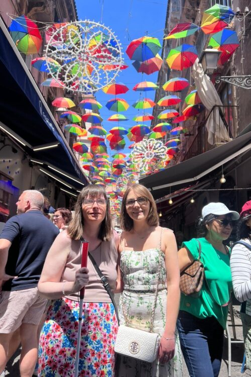 Holly and Elin standing under a canopy of bright striped umbrellas on a narrow street, smiling at the camera. Above them, dozens of small umbrellas in red, yellow, green, and blue are suspended in rows, creating a vibrant ceiling against a clear blue sky. Holly wears glasses, a sleeveless beige crop top, a colourful floral patterned skirt and a black crossbody bag. Elin wears glasses and a sleeveless dress with a green floral pattern, and a white crossbody bag.
