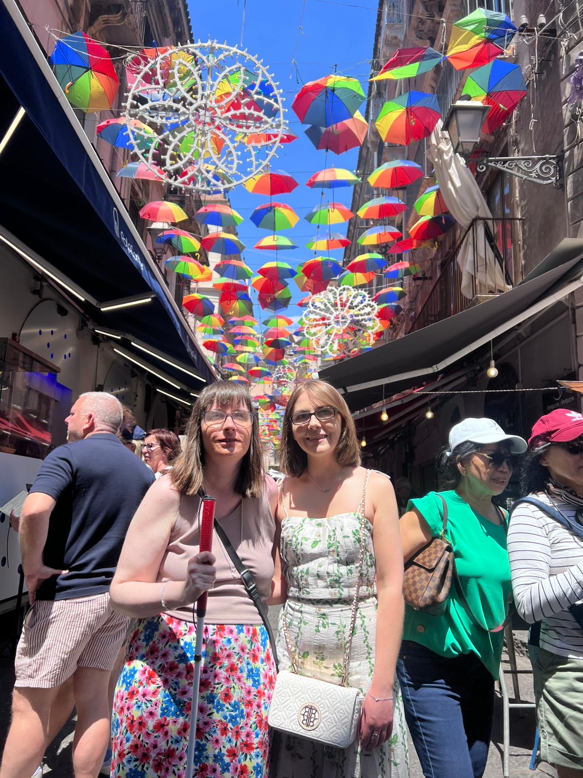 Holly and Elin standing under a canopy of bright striped umbrellas on a narrow street, smiling at the camera. Above them, dozens of small umbrellas in red, yellow, green, and blue are suspended in rows, creating a vibrant ceiling against a clear blue sky. Holly wears glasses, a sleeveless beige crop top, a colourful floral patterned skirt and a black crossbody bag. Elin wears glasses and a sleeveless dress with a green floral pattern, and a white crossbody bag.