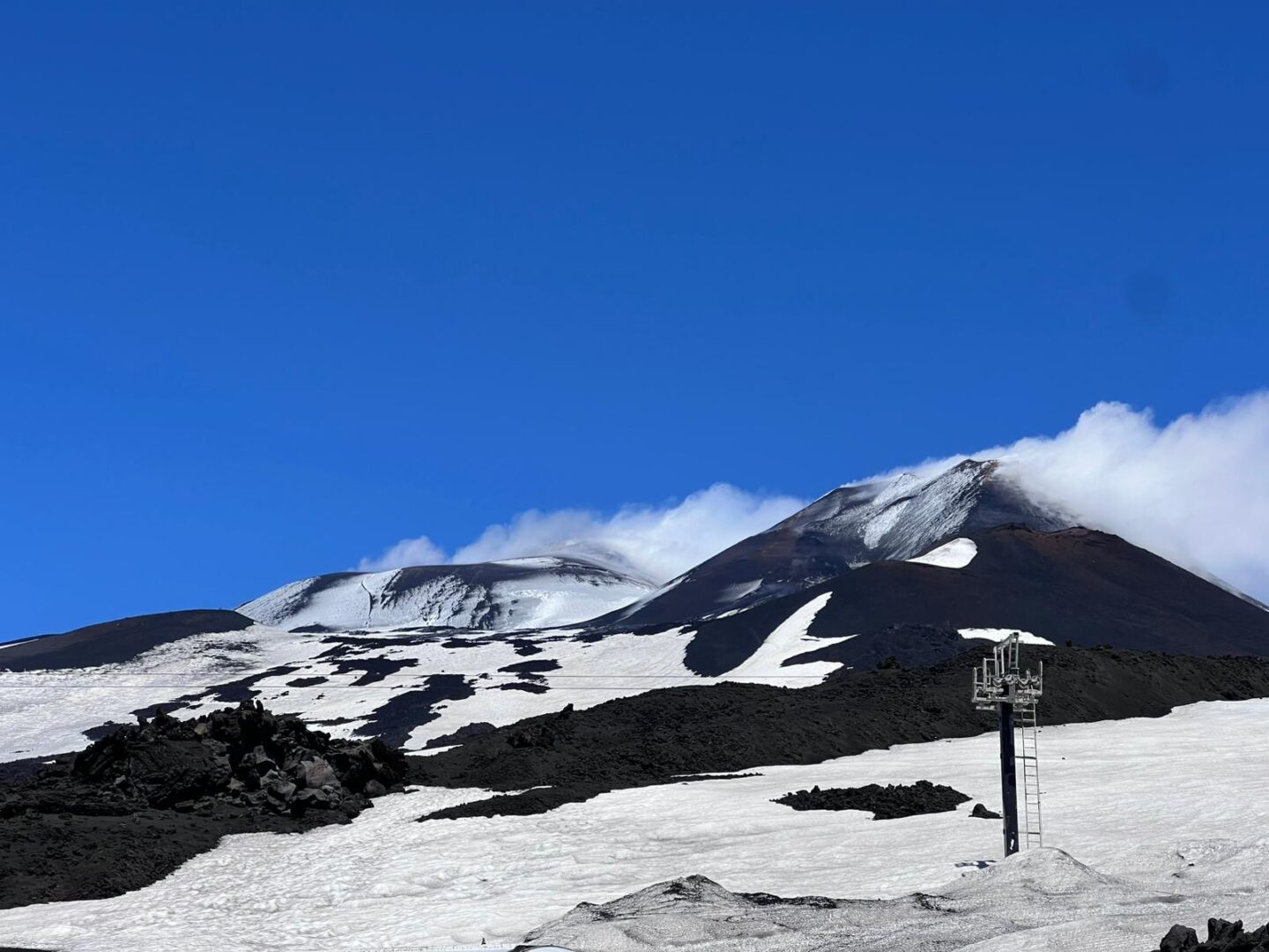 A snow-capped Mount Etna under a deep blue sky. The mountain is so high, the peak appears within the clouds. In the foreground, there is a pylon for the cable car. Black volcanic rock can be seen breaking out from the snow.